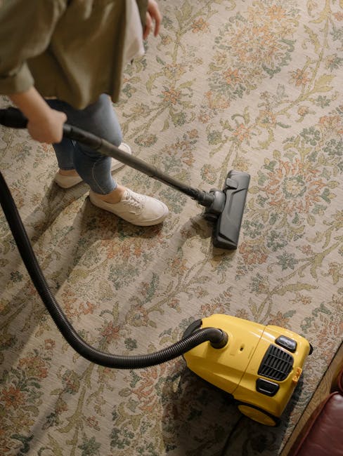 Close-up of a professional vacuum cleaner with a transparent dust container filled with debris, connected to a flexible hose with a patterned surface. The vacuum is positioned on a plush, pinkish-beig